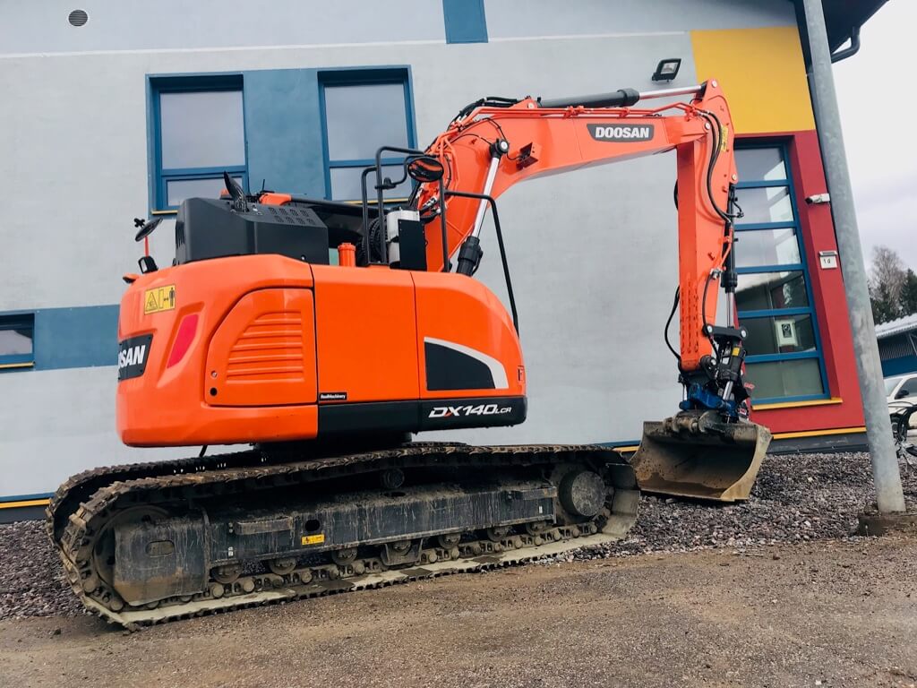 Oranssi Doosan DX140LCR -kaivinkone rakennuksen edustalla, jossa sinisiä ja keltaisia ikkunakehyksiä.