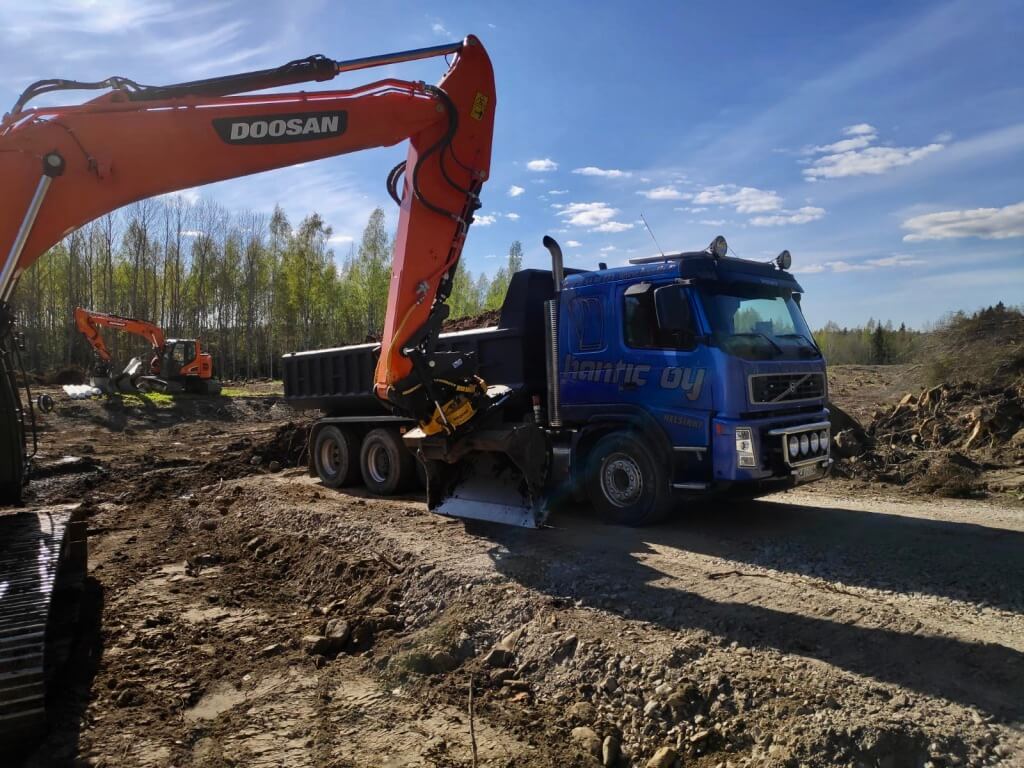 Sininen Volvo-dumpperi ja oranssi Doosan-kaivinkone työmaalla, metsän reunassa aurinkoisena päivänä.