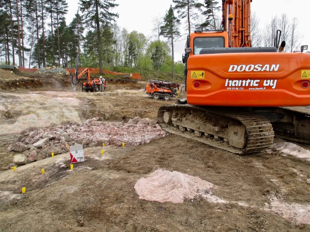 Rakennustyömaa metsäisellä alueella, jossa näkyy oranssi Doosan-kaivuri ja muita rakennuskoneita.