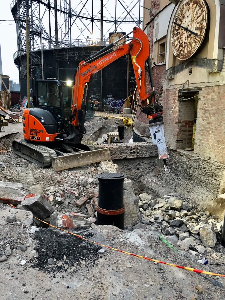 Oranssi Hitachi-kaivinkone työmaalla murskaamassa betonia rakennuksen vieressä, taustalla suuri teräsrakenne ja ruosteinen kello seinässä.