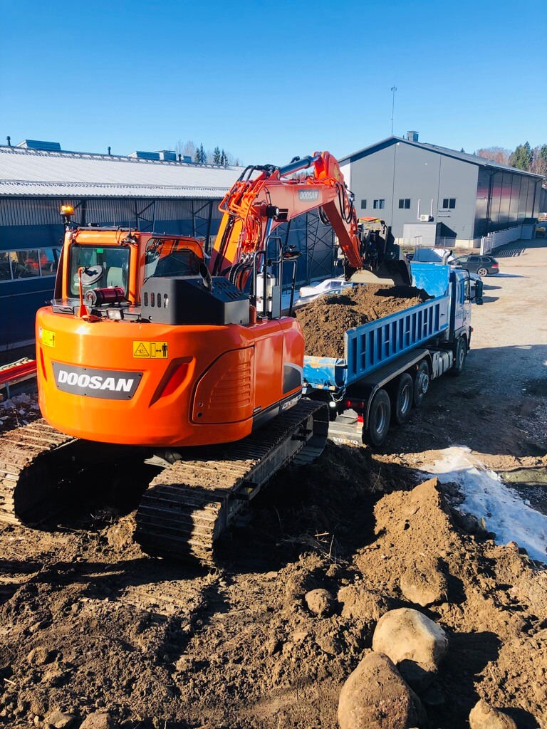 Oranssi Doosan-kaivinkone lastaa maata siniseen rakennustyömaan rekkaan aurinkoisena päivänä.