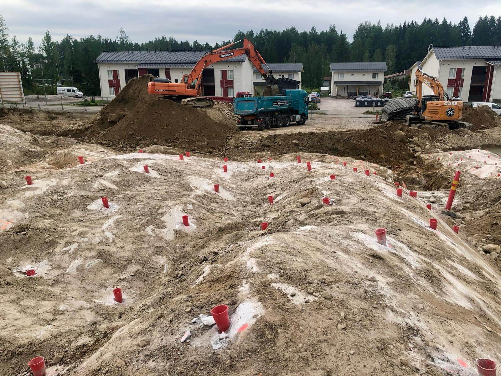 Rakennustyömaa, jossa kaksi kaivinkonetta lastaamassa maata kuorma-autoon asuinrakennusten ja metsän edustalla.