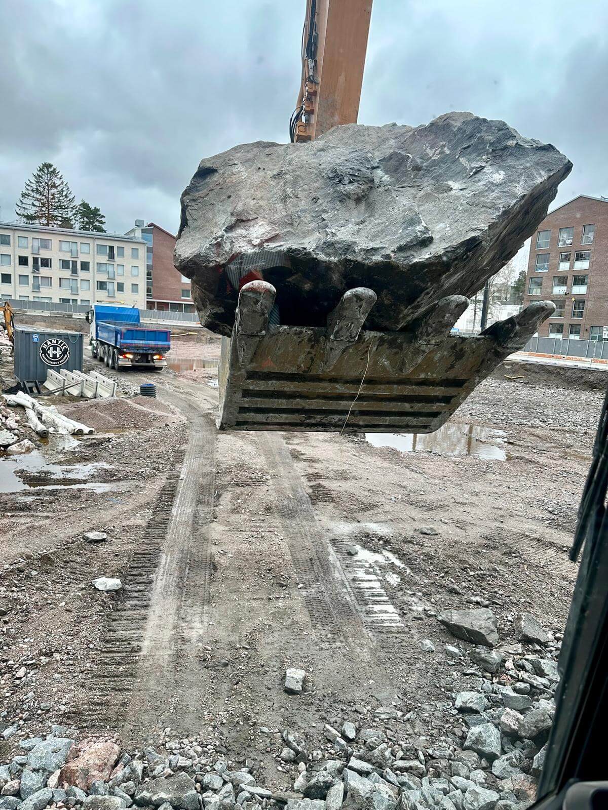 Kaivinkoneen kauha nostaa suuren kallion rakennustyömaalla, taustalla rakennuksia ja kuorma-auto.