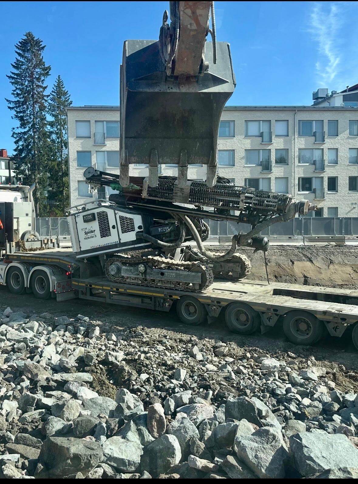 Maastossa kivetty alue, jossa rakennustyömaalla matalalla oleva kaivinkone ja suuri kaivinkoneen kauha.