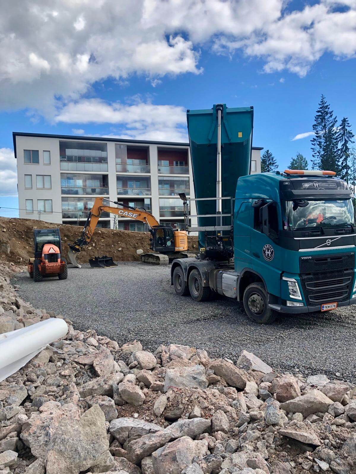 Kuorma-auto, kaivinkone ja pieni telakone rakennustyömaalla kerrostalon edustalla.