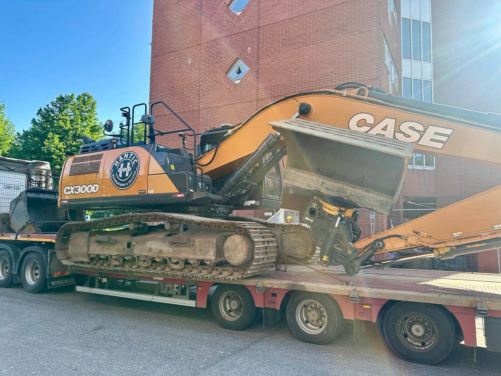 Kaivinkone CX300D, jossa Hantic-merkkinen logo, kuljetuksen aikana kuorma-auton perävaunulla kerrostalon edessä.