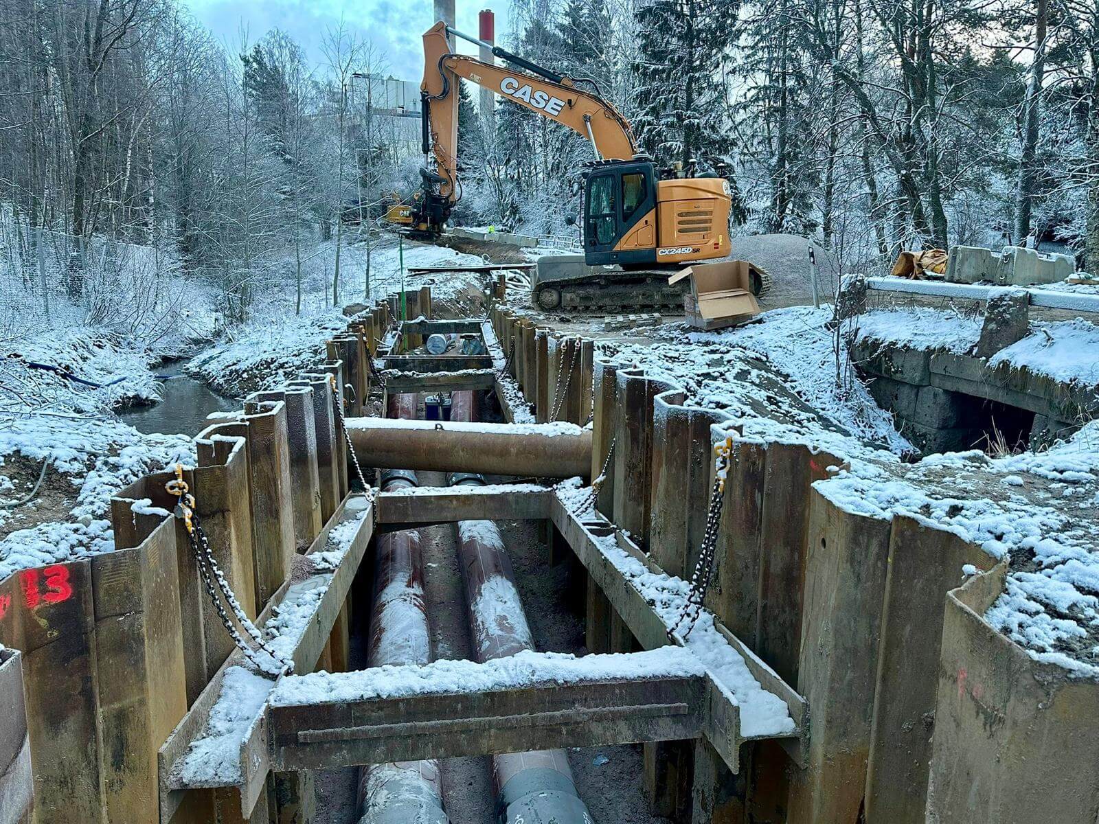Lumisessa metsämaisemassa talvella työmaa, jossa kaivinkone ja maanalaisia putkia ympäröivillä teräksisillä tuentarakenteilla.