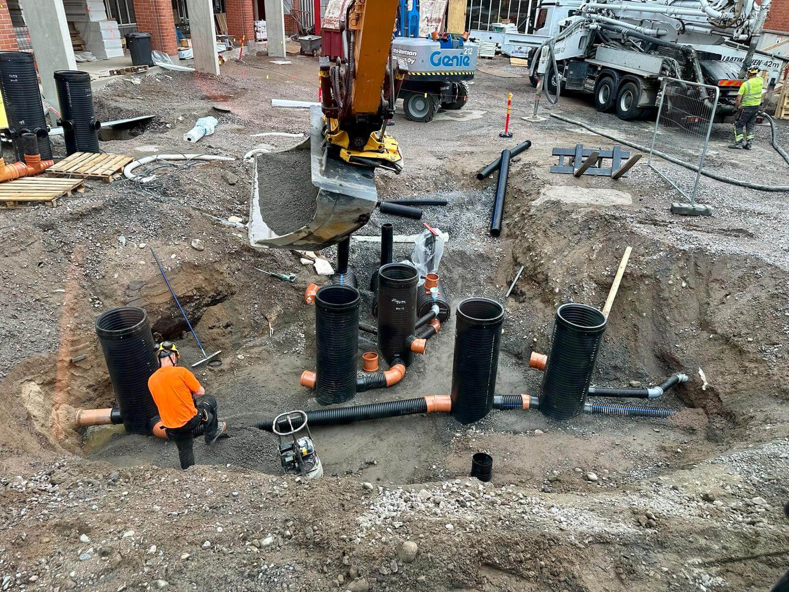 Rakennustyömaa, jossa työntekijä oranssissa paidassa asentaa mustia putkia kaivannon pohjalle kaivinkoneen kauhan alla.