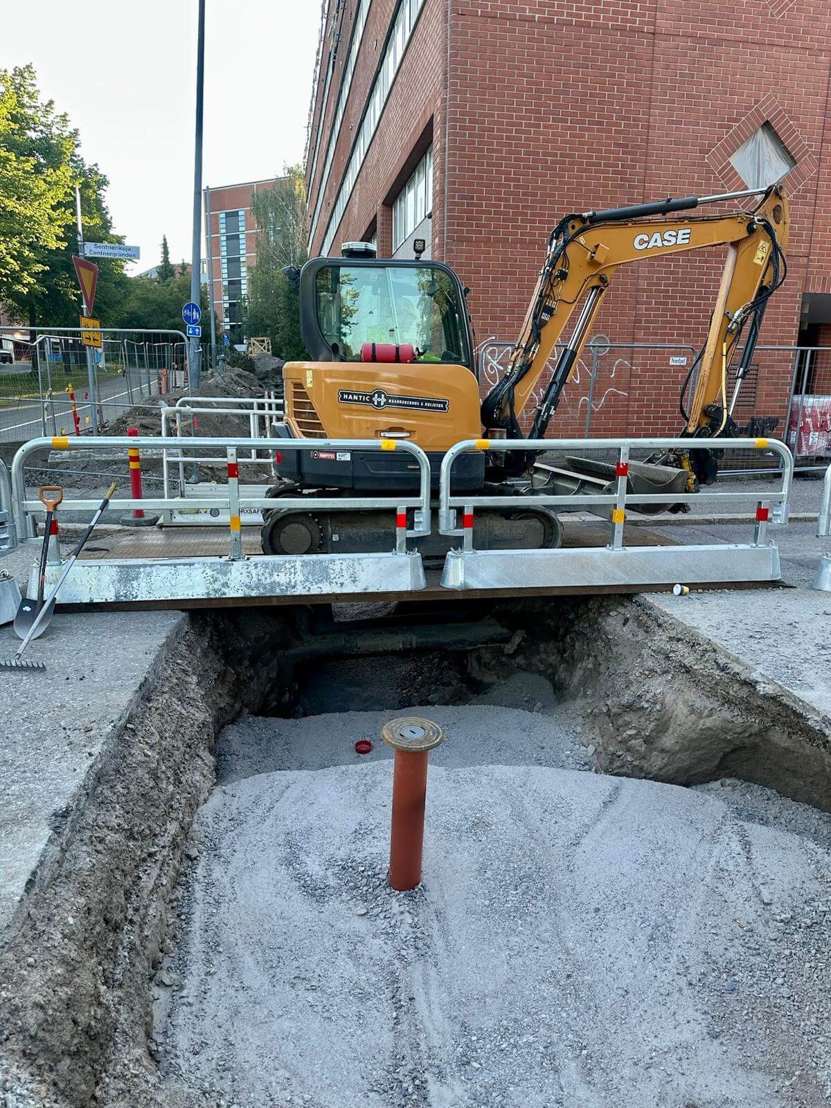 Rakennustyömaa kadulla, jossa on kaivuri ja kaivettu alue betoniseinää vasten.