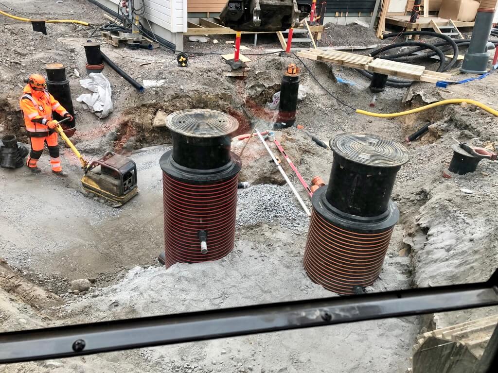 Rakennustyöntekijä työskentelee keltaisella maarakennuskoneella rakennustyömaalla, jossa on kaivettu maata ja näkyvissä suuria mustia putkia ja rakennusmateriaaleja.