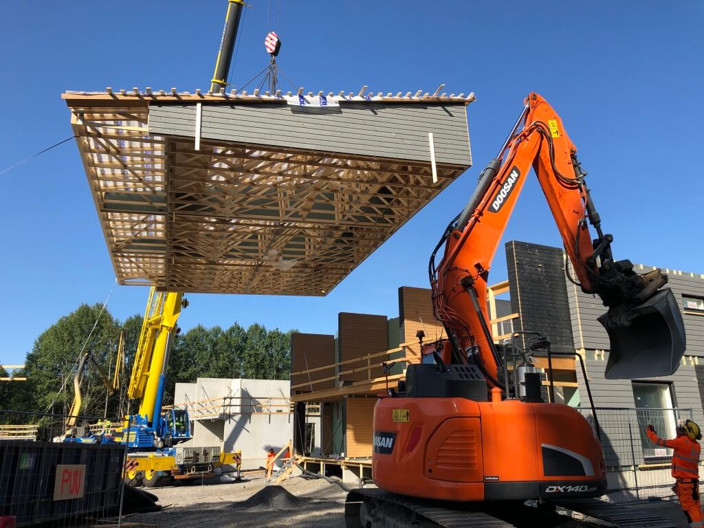 Rakennustyömaalla nosturi nostaa puurakenteista rakennuksen osaa paikoilleen, vieressä oranssi Doosan-kaivinkone.