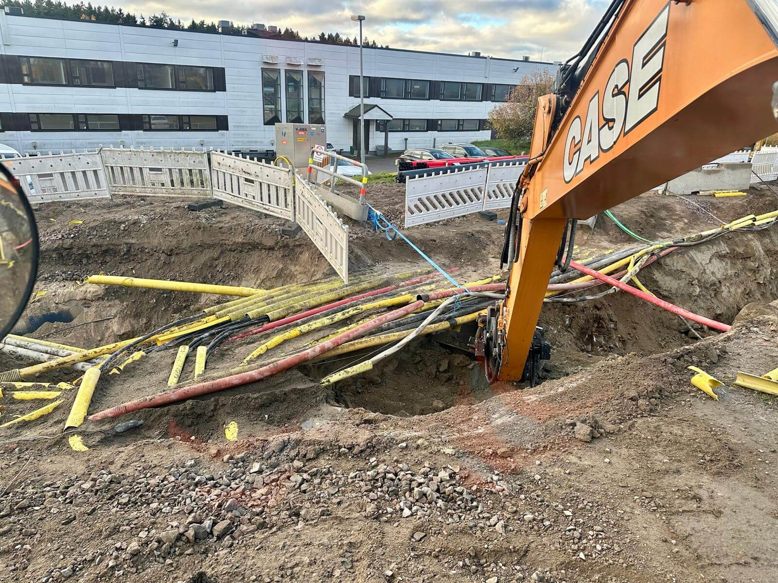 Kaivinkone työmaalla, maassa kaapeleita ja putkia, taustalla toimistorakennus ja autoja.