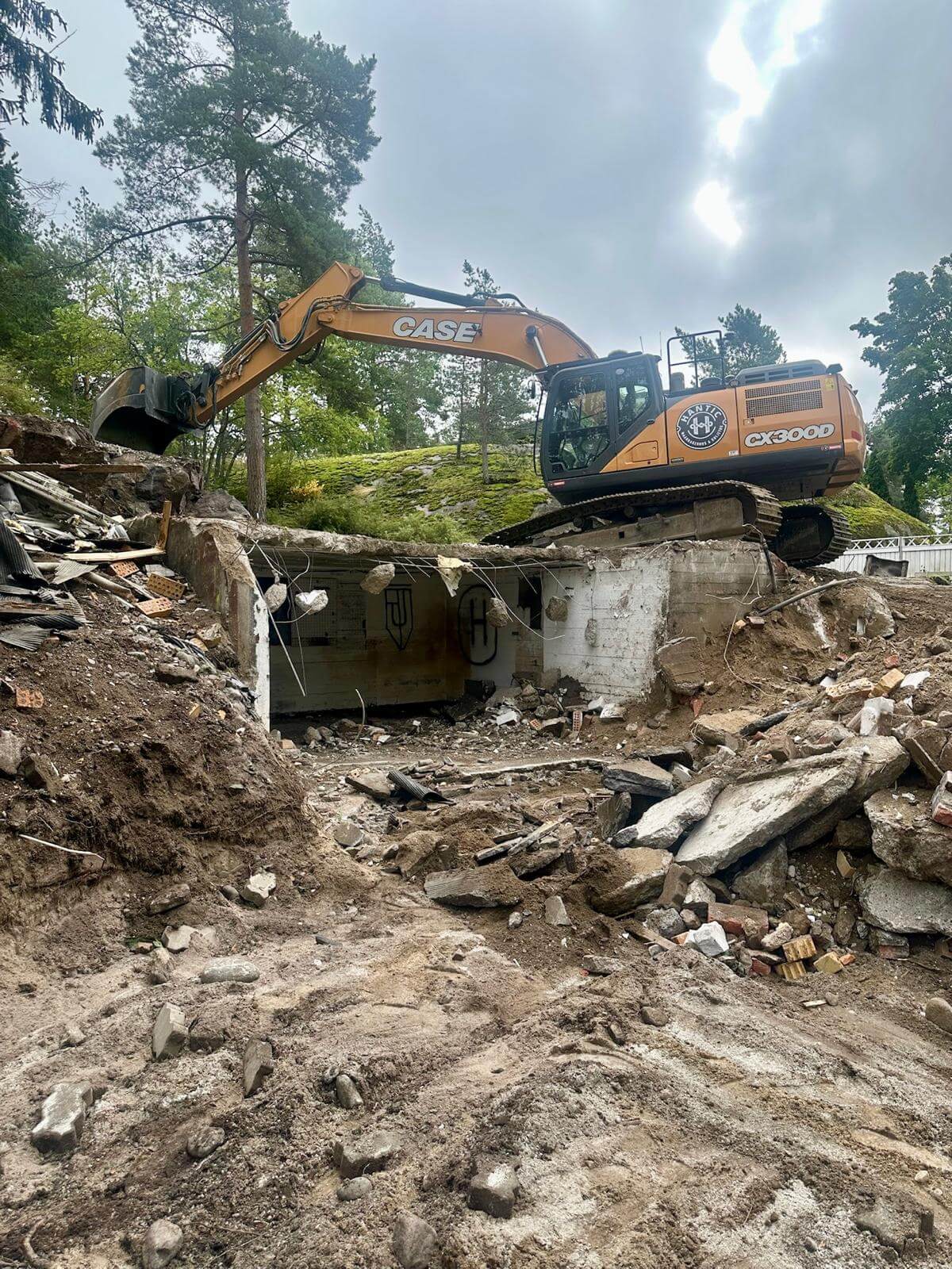Kaivinkone purkamassa rapistunutta betonirakennusta metsäisessä ympäristössä.