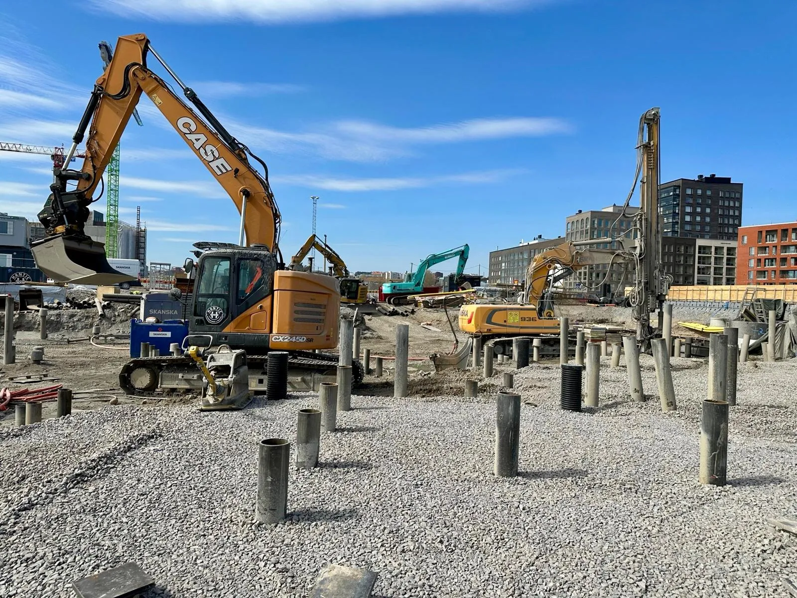 Rakennustyömaa, jossa kaksi kaivinkonetta ja betoniputkia kivikentällä, taustalla moderneja rakennuksia ja sininen taivas.