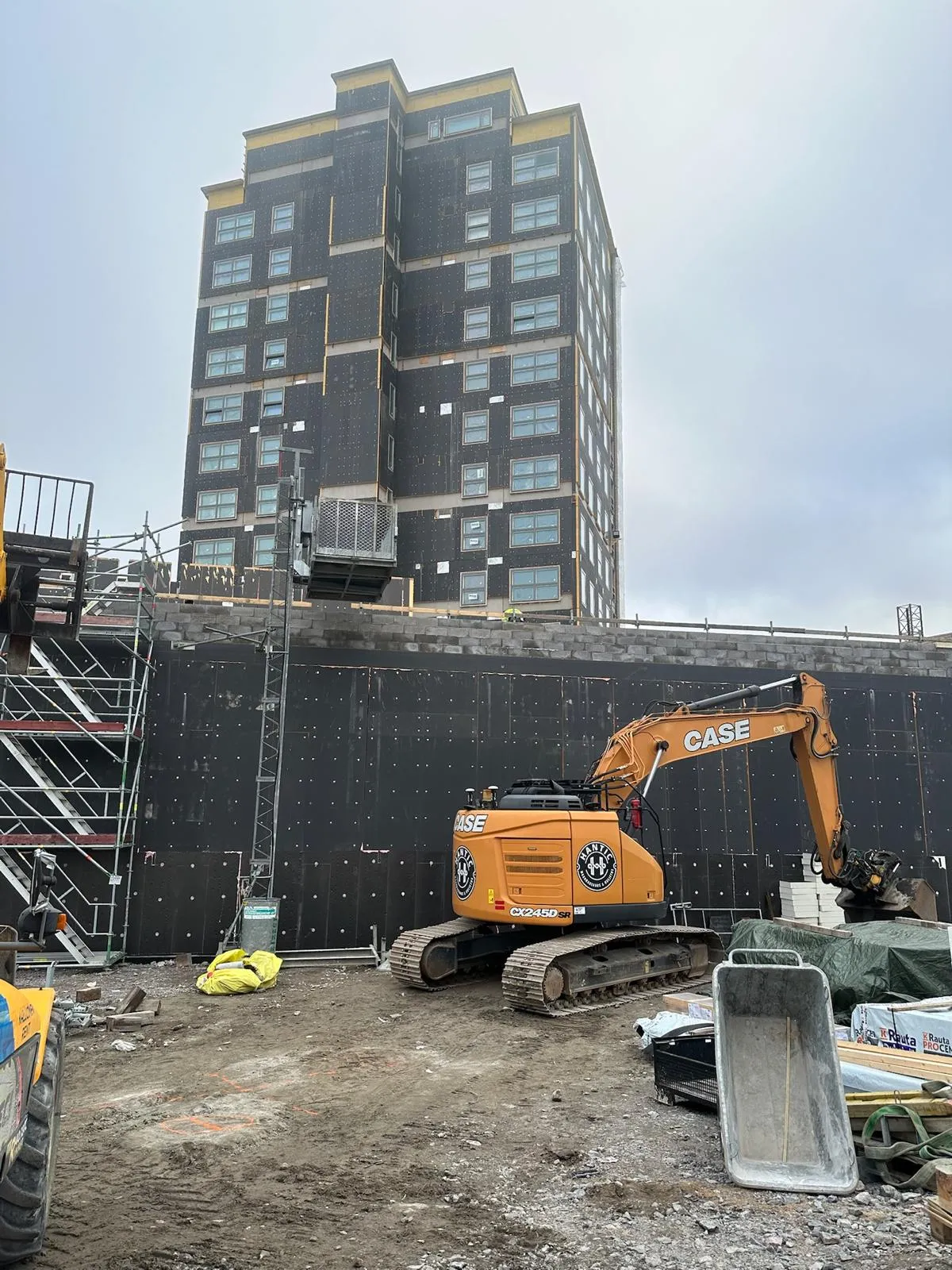 Rakennustyömaa, jossa oranssi kaivinkone CASE ja musta rakennus taustalla.