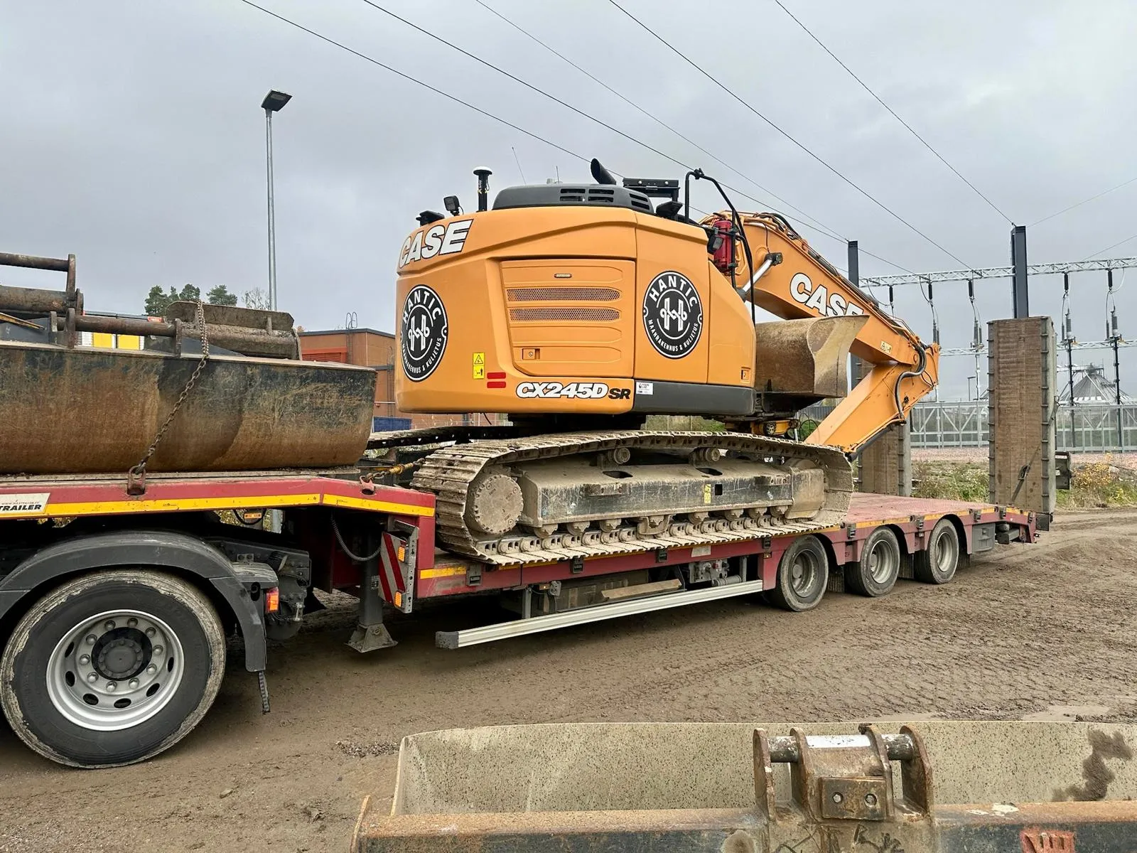 Oranssi kaivinkone on kuorma-autotrailerin päällä teollisuusalueella pilvisenä päivänä.