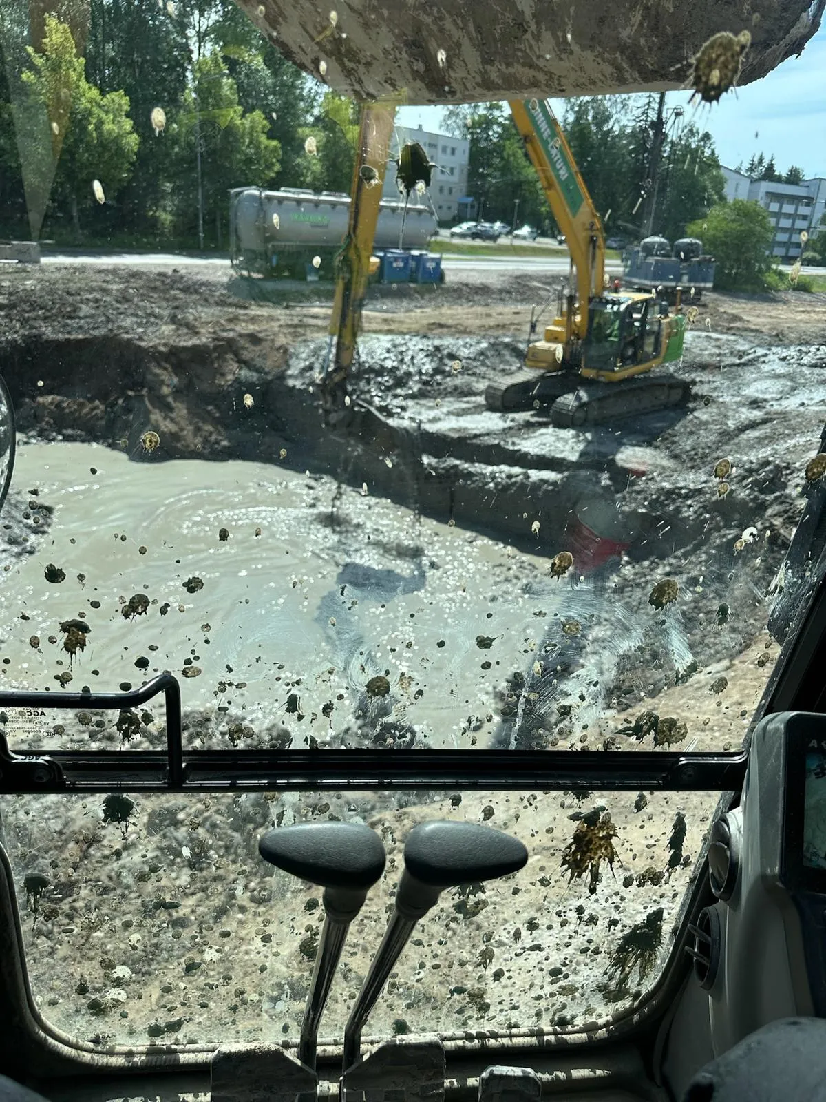 Likainen kaivinkoneen ohjaamon ikkuna, josta näkyy toinen kaivinkone kaivamassa mutaista maata rakennustyömaalla.