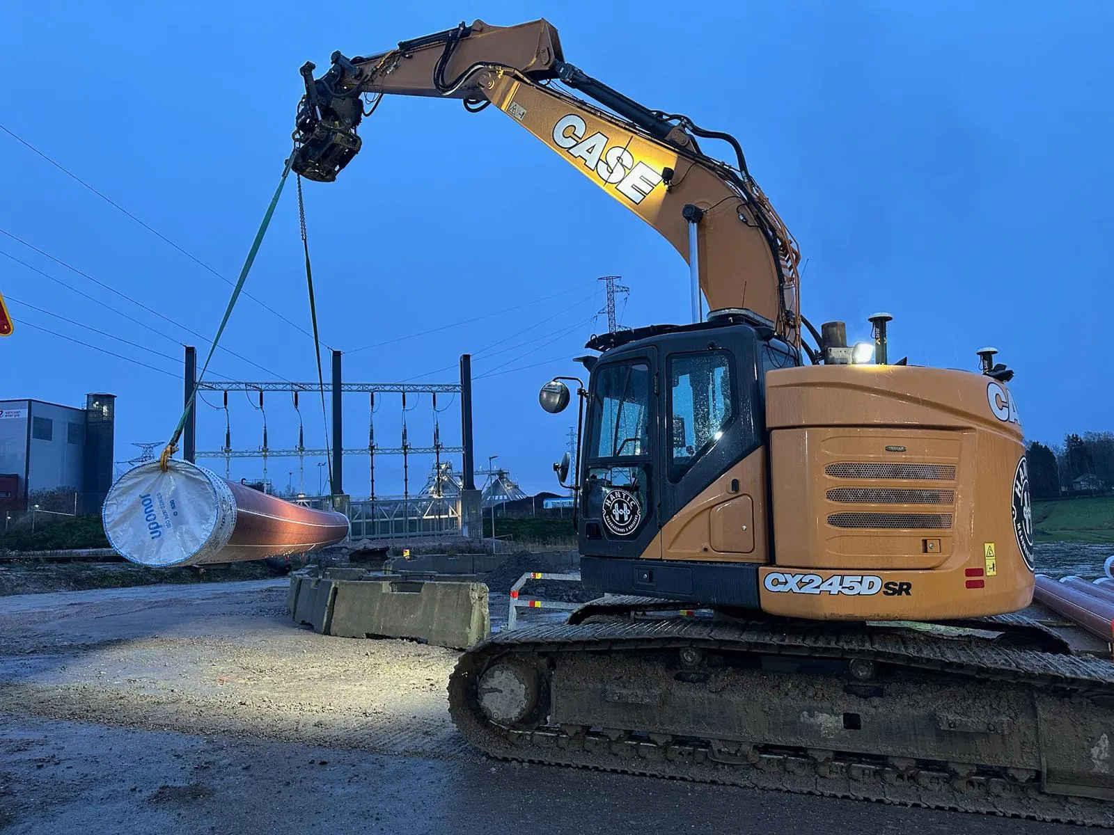 Kaivinkone nostaa pitkää putkea työmaalla hämärässä valaistuksessa.