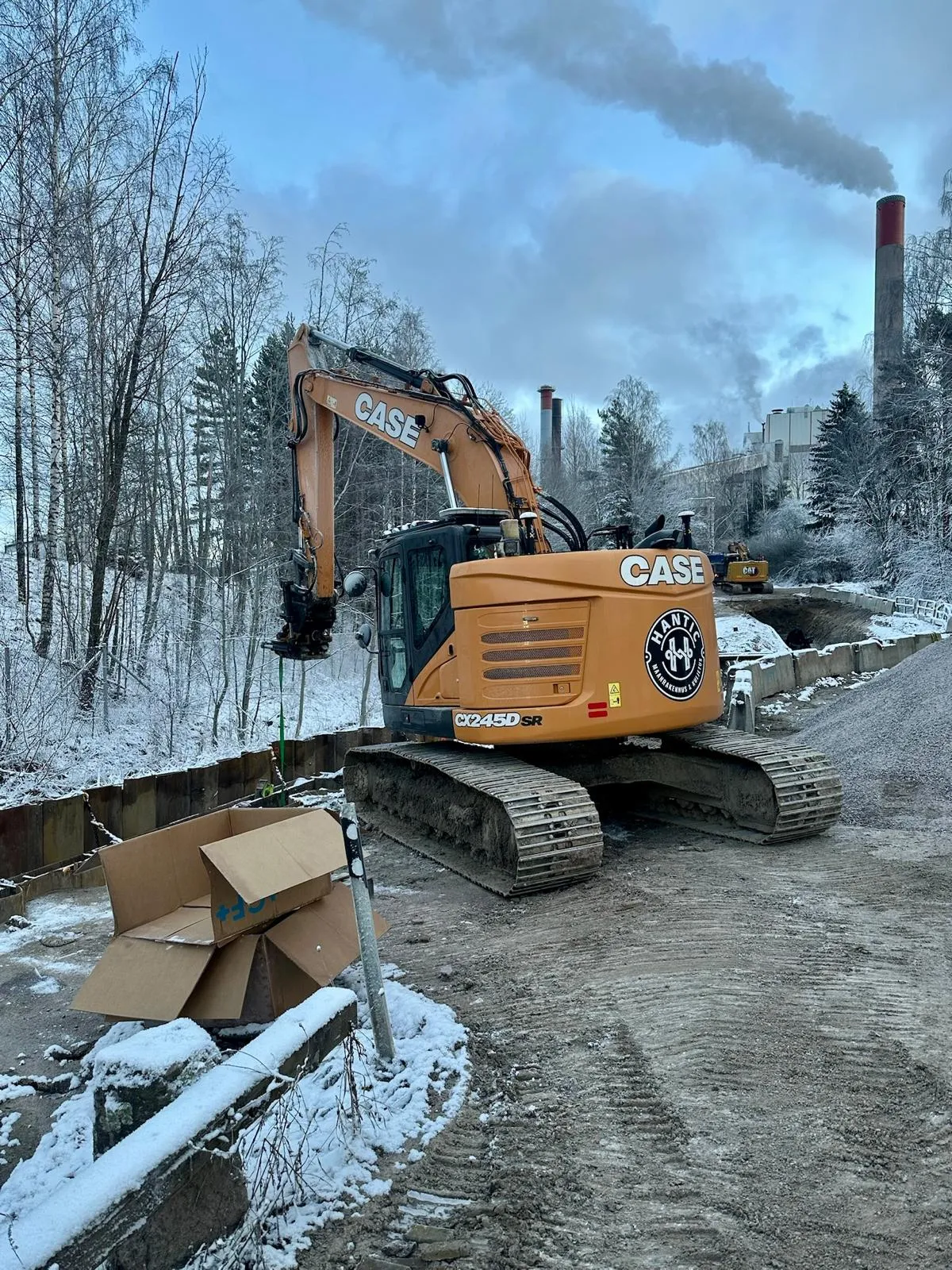 Oranssi CASE-kaivinkone työmaalla talvisessa maisemassa, lumisia puita ja savua nousee tehtaasta taustalla.