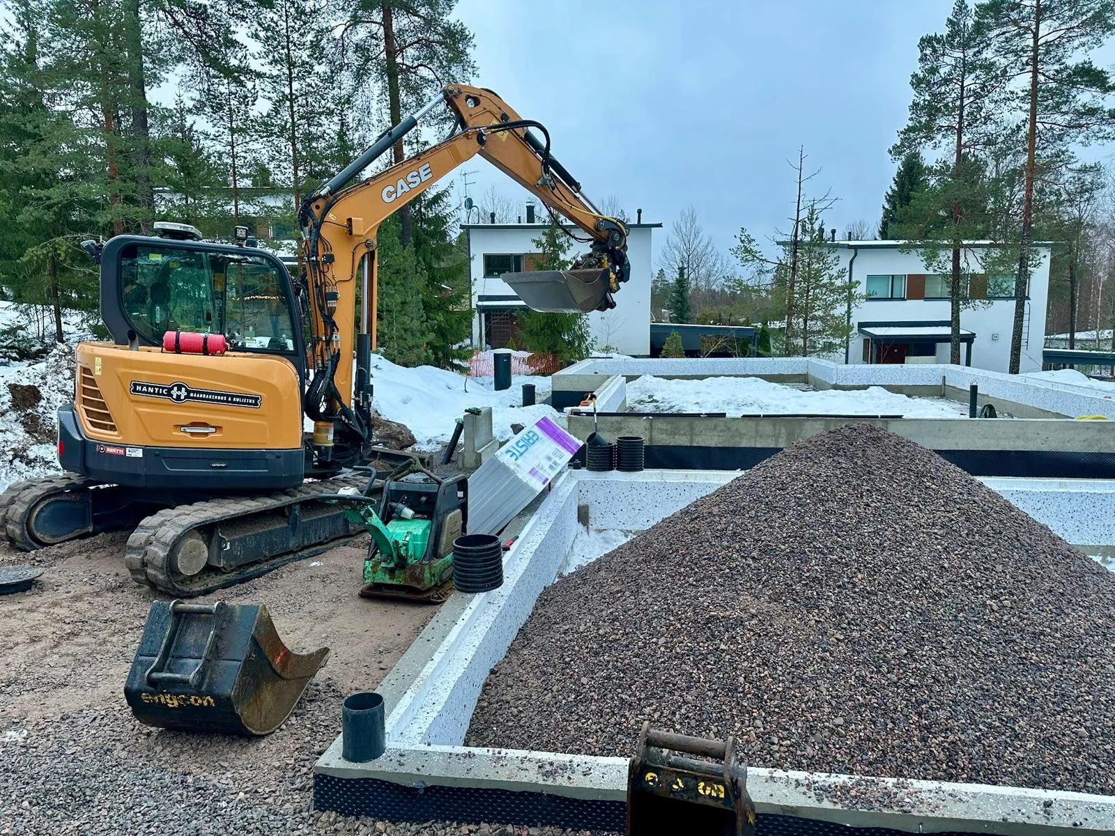 Rakennustyömaa, jossa keltainen CASE-kaivinkone ja suuri sorakasa perustuksen sisällä.