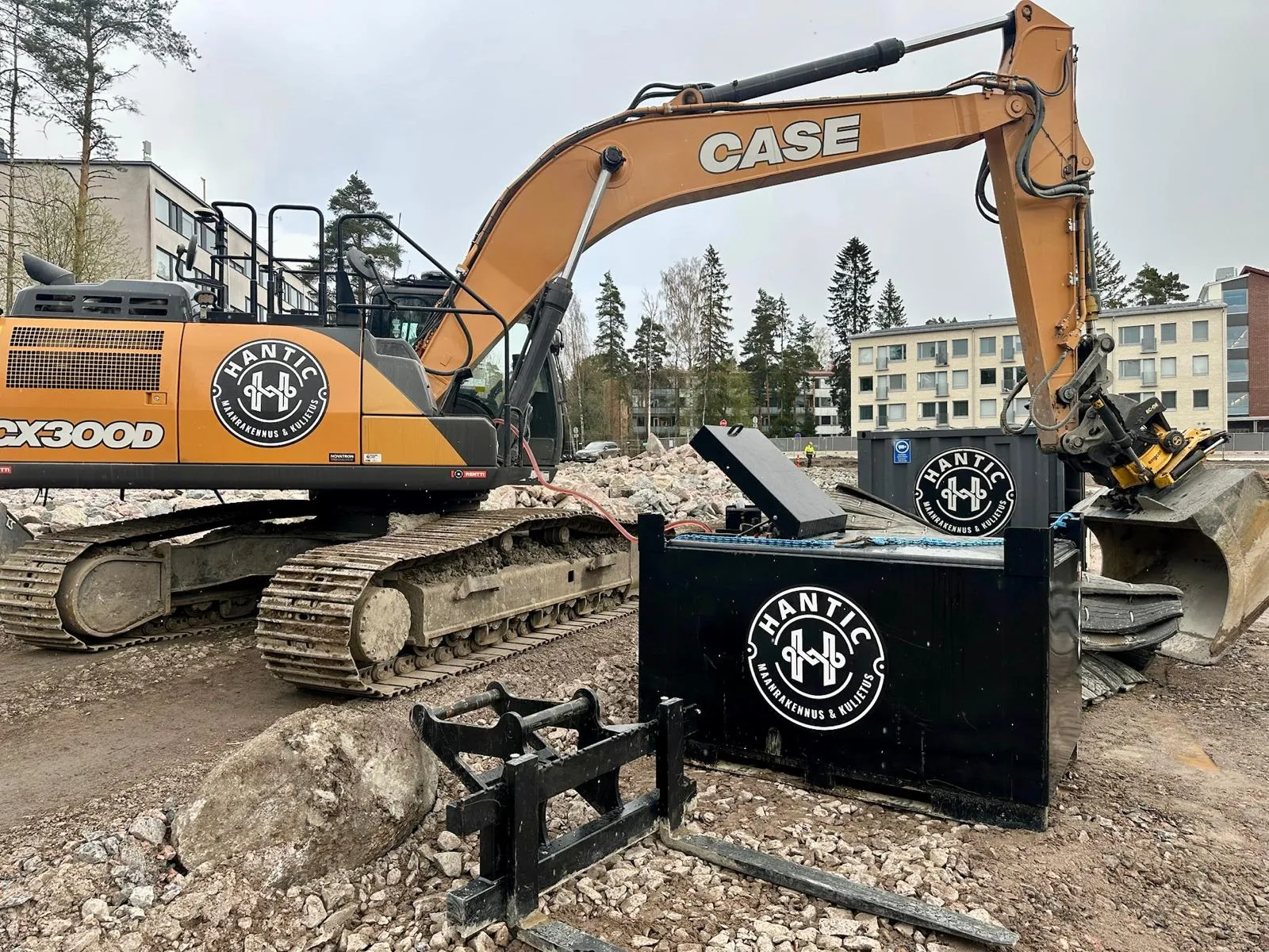 Rakennustyömaan kaivinkone, jossa on Hantic-yhtiön logo ja rakennuksien tausta.