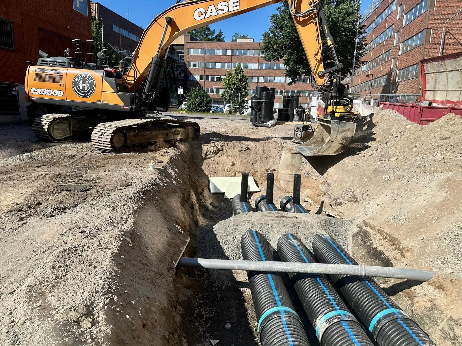Rakennustyömaa, jossa iso kaivinkone ja maahan asennettuja suuria mustia muoviputkia.