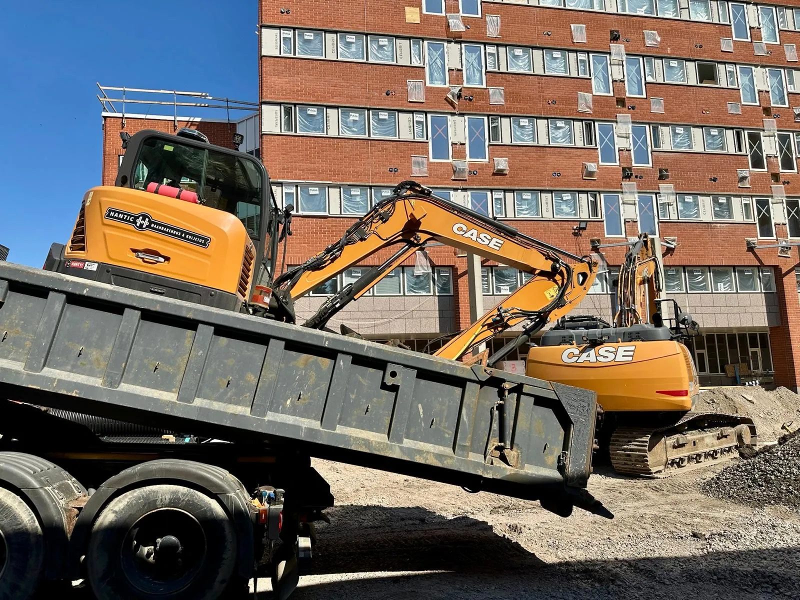 Keltainen CASE-kaivinkone työmaalla punatiilisen rakennuksen edessä kirkkaalla säällä.