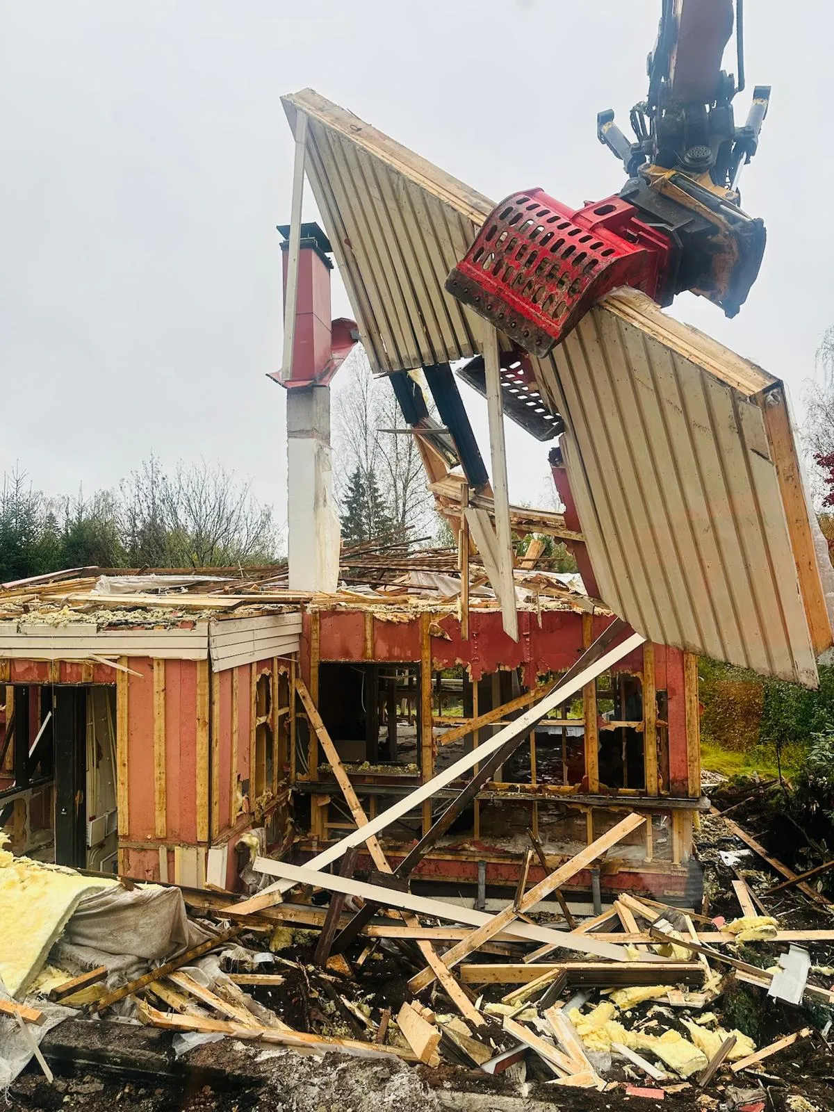 Rakennus puretaan, ja suuri punainen kaivinkoneen kiinnike nostaa rakennuksen kattopaneelin pois.