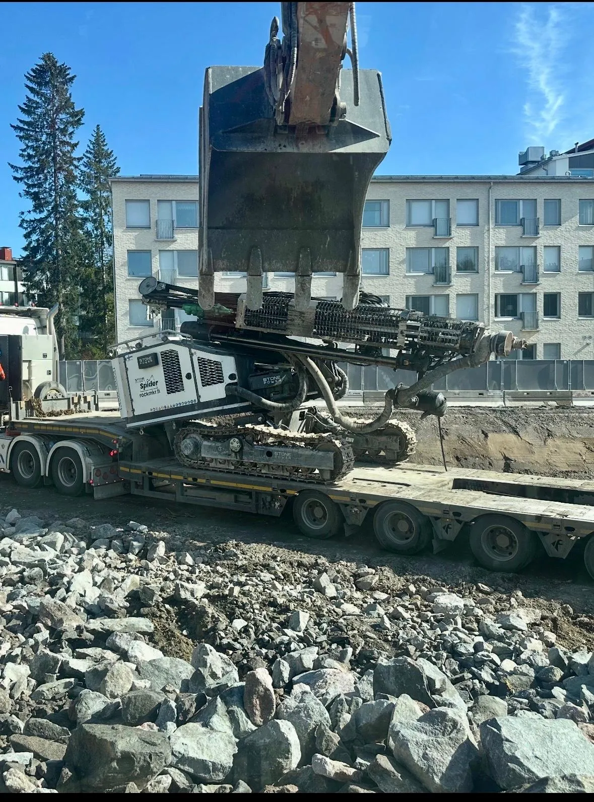 Kaivinkoneen kauha nousee ylös kuorma-auton lavalla olevasta kaivoskoneesta rakennustyömaalla, taustalla asuinrakennus ja kirkas sininen taivas.