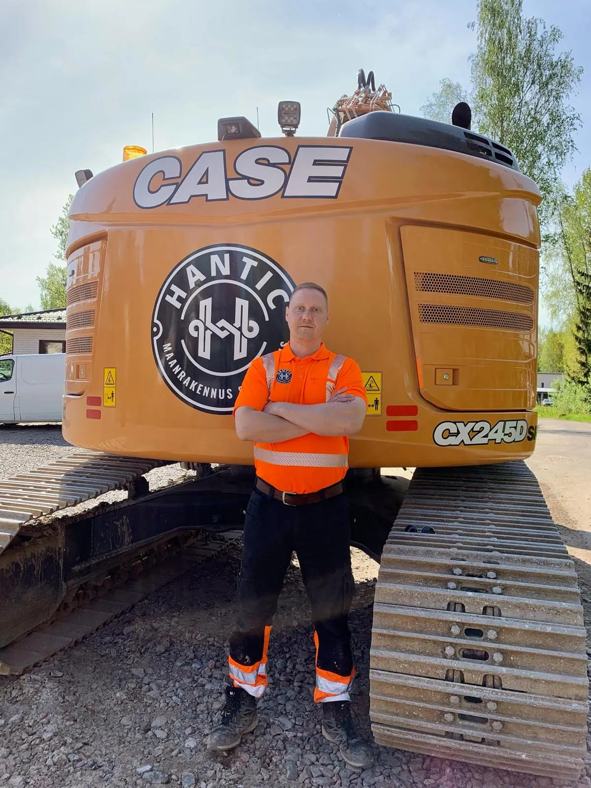 Mies oranssin näkyvyyspaidan ja mustien työhousujen kanssa seisoo ristiin asetetuin käsivarsin suuren keltaisen kaivinkoneen edessä.