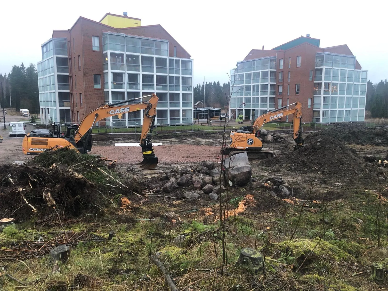 Kaksi oranssia kaivinkonetta työskentelee rakennustyömaalla kahden punatiilisen talon edessä sumuisena päivänä.
