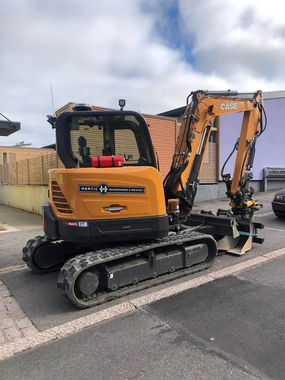 Keltainen kaivinkone, jonka kyljessä on Hantic Maanrakennus & Kuljetus -teksti, pysäköity kadun reunalle.