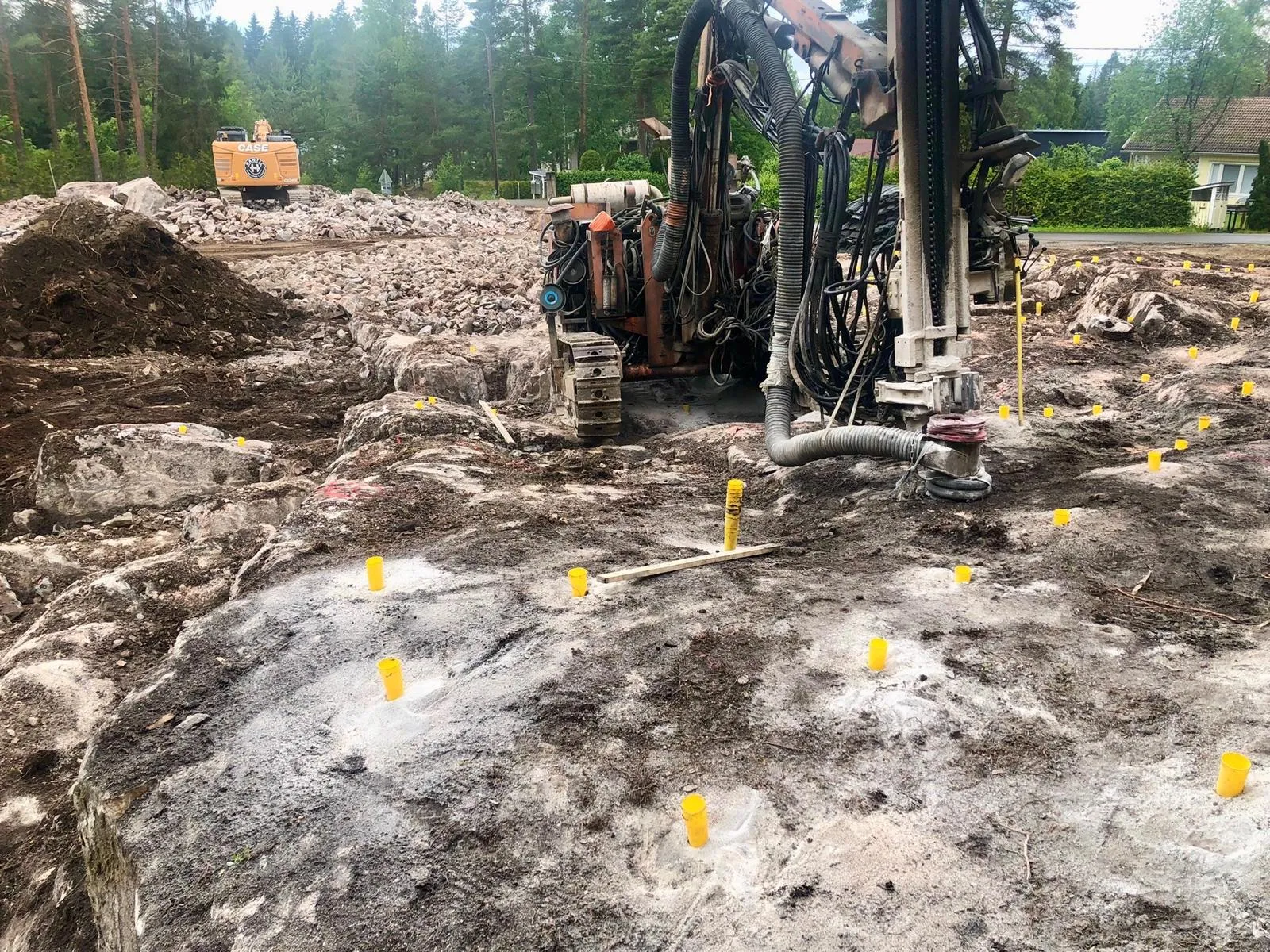 Rakennustyömaa, jossa louhittu kalliopinta ja keltaisia merkintäpisteitä sekä kaivinkone ja porauskone metsäisen taustan edessä.