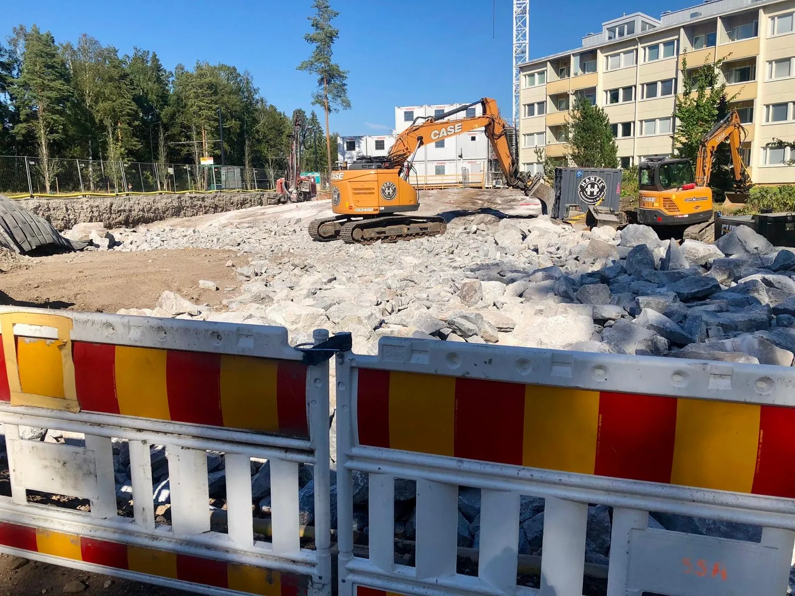 Rakennustyömaa aidatulla alueella, jossa kaksi oranssia kaivinkonetta ja suuret kiviainekset edustalla.