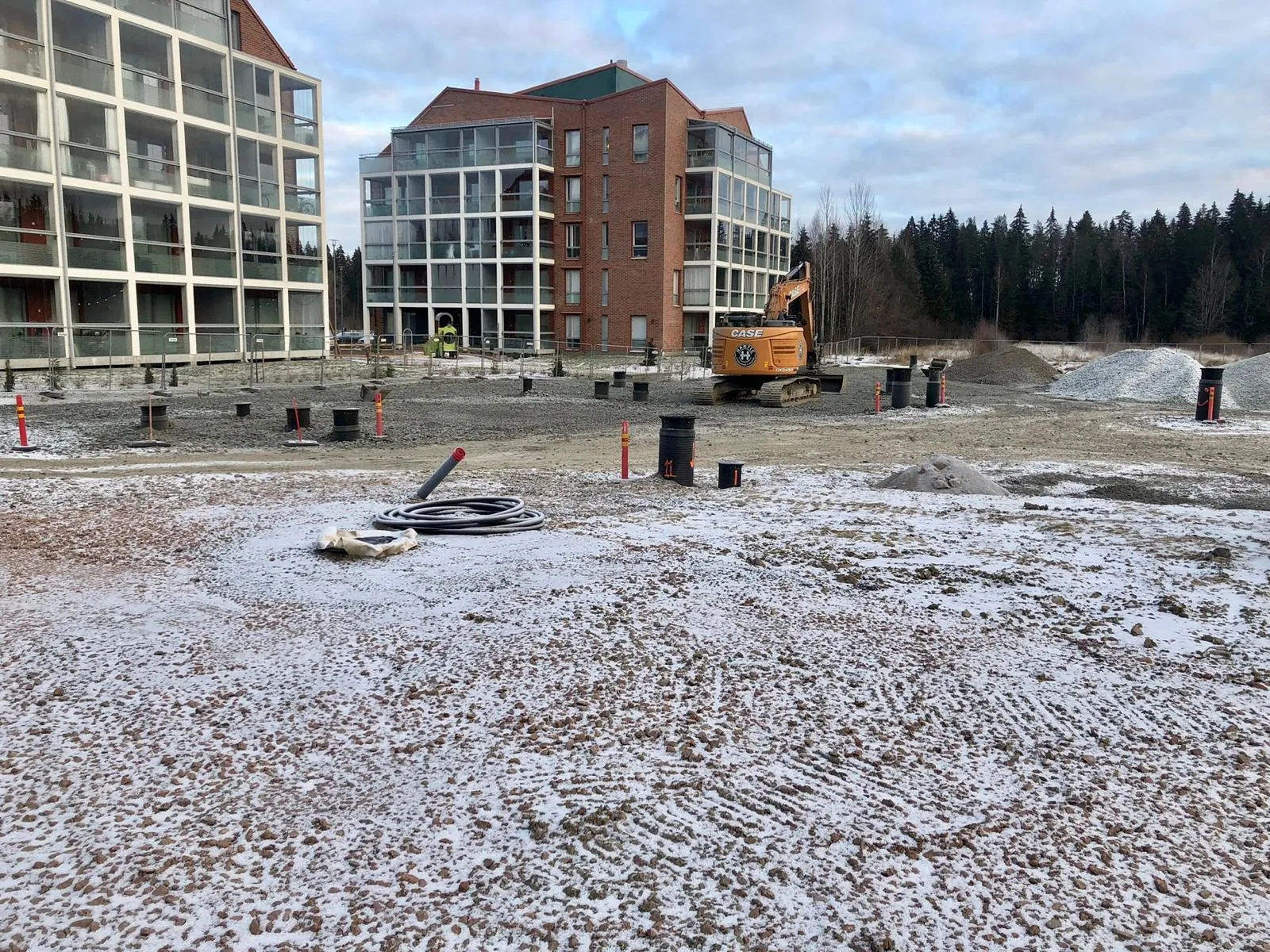 Rakennustyömaaalue, jossa näkyy hiekoitettu maapohja, kaivinkone ja modernit monitorniset asuinrakennukset taustalla.